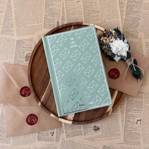 K&ouml;nnte beinhalten: Ein Buch mit dem Titel "The Last Letter" mit mintgr&uuml;nem Umschlag und Umschlagillustrationen, auf einem Holztablett. Zwei versiegelte Umschl&auml;ge und ein kleiner Strau&szlig; Trockenblumen sind ebenfalls vorhanden, alles vor dem Hintergrund alter Zeitungsseiten.