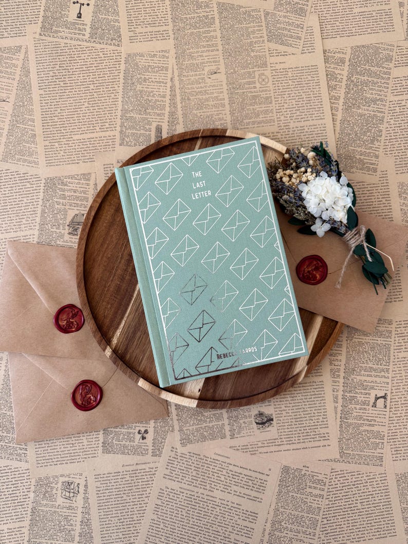 K&ouml;nnte beinhalten: Ein Buch mit dem Titel "The Last Letter" mit mintgr&uuml;nem Umschlag und Umschlagillustrationen, auf einem Holztablett. Zwei versiegelte Umschl&auml;ge und ein kleiner Strau&szlig; Trockenblumen sind ebenfalls vorhanden, alles vor dem Hintergrund alter Zeitungsseiten.