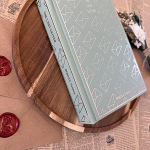 K&ouml;nnte beinhalten: Ein Hardcover-Buch mit dem Titel "The Last Letter" von Rebecca Yarros, mit hellt&uuml;rkisfarbenem Einband und silbernem Umschlagmuster. Das Buch liegt auf einem Holztablett mit braun-goldener Oberfl&auml;che, zusammen mit einem versiegelten Umschlag und Trockenblumen.