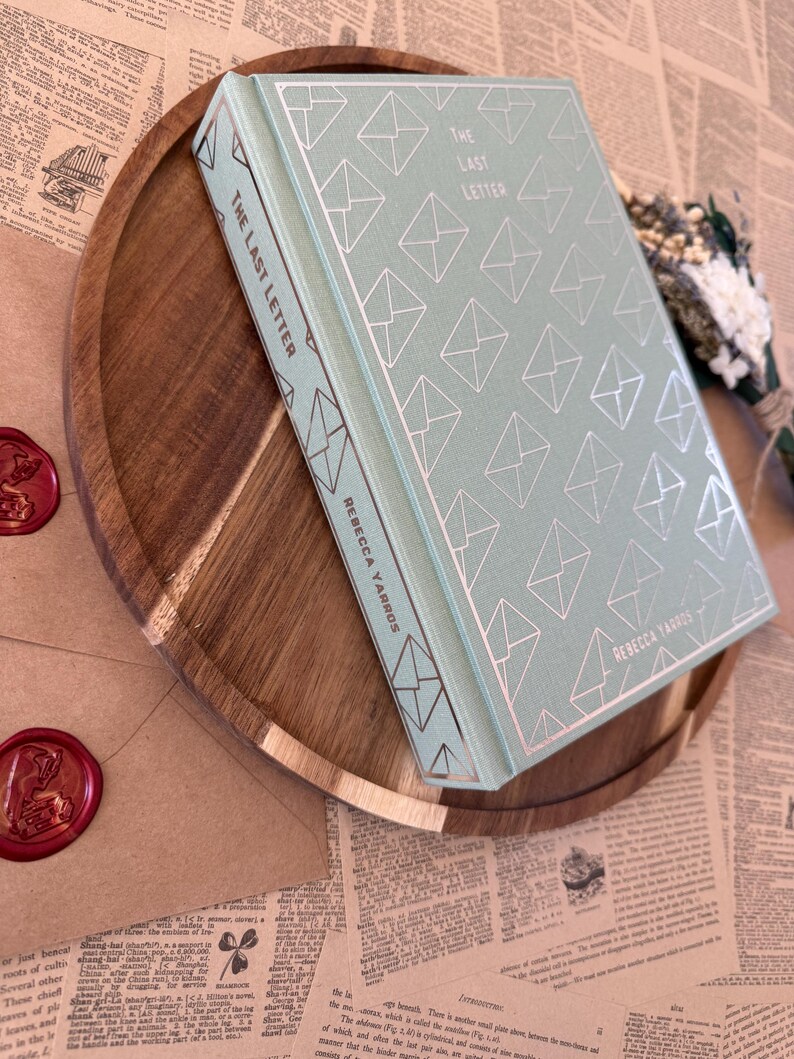 K&ouml;nnte beinhalten: Ein Hardcover-Buch mit dem Titel "The Last Letter" von Rebecca Yarros, mit hellt&uuml;rkisfarbenem Einband und silbernem Umschlagmuster. Das Buch liegt auf einem Holztablett mit braun-goldener Oberfl&auml;che, zusammen mit einem versiegelten Umschlag und Trockenblumen.