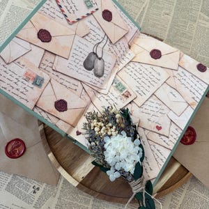K&ouml;nnte beinhalten: Ein dekoratives offenes Buch mit Vintage-Briefdesign, Briefmarken und Wachssiegeln. Ein kleiner Strau&szlig; Trockenblumen und ein Paar Erkennungsmarken liegen auf einem Holztablett, umgeben von bedrucktem Papier.