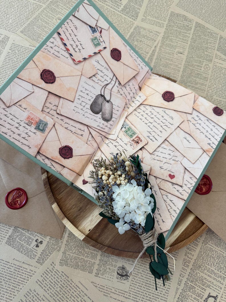 K&ouml;nnte beinhalten: Ein dekoratives offenes Buch mit Vintage-Briefdesign, Briefmarken und Wachssiegeln. Ein kleiner Strau&szlig; Trockenblumen und ein Paar Erkennungsmarken liegen auf einem Holztablett, umgeben von bedrucktem Papier.