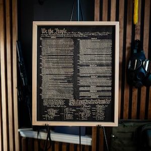 K&ouml;nnte beinhalten: Ein gerahmter Druck der Verfassung der Vereinigten Staaten auf einem Holzgrund. Der Text ist in Schwarzwei&szlig; und lautet "We the People".