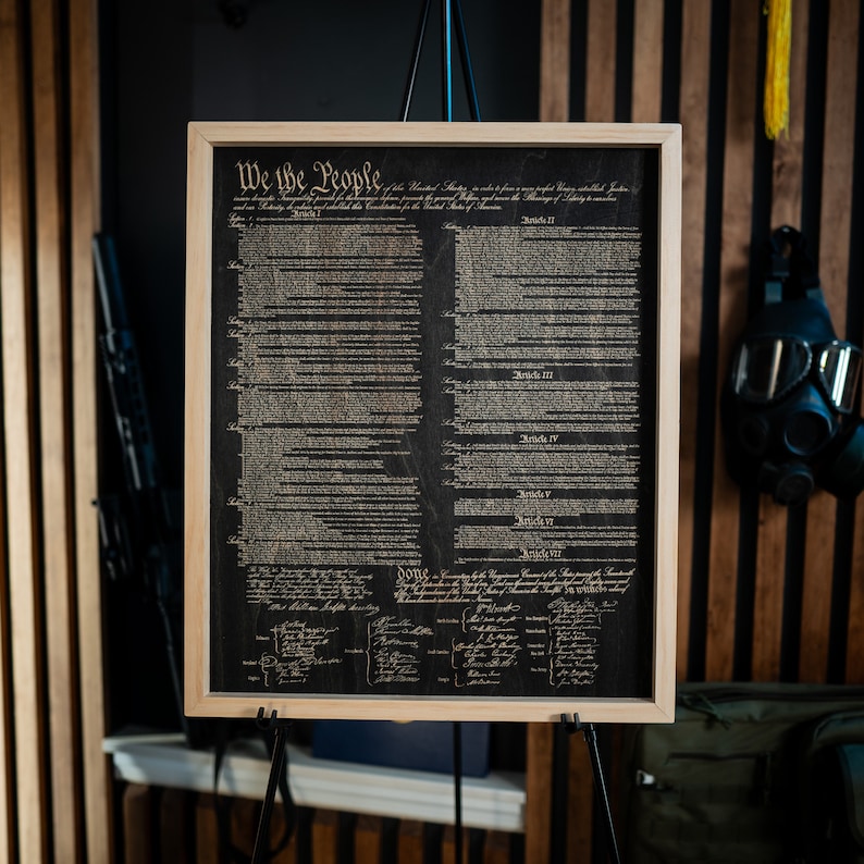 K&ouml;nnte beinhalten: Ein gerahmter Druck der Verfassung der Vereinigten Staaten auf einem Holzgrund. Der Text ist in Schwarzwei&szlig; und lautet "We the People".