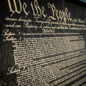 K&ouml;nnte beinhalten: Eine Nahaufnahme eines gerahmten Dokuments mit dem Text "We the People" und den ersten Abschnitten der Verfassung der Vereinigten Staaten. Das Dokument ist auf einem dunklen Hintergrund mit wei&szlig;em Text gedruckt.