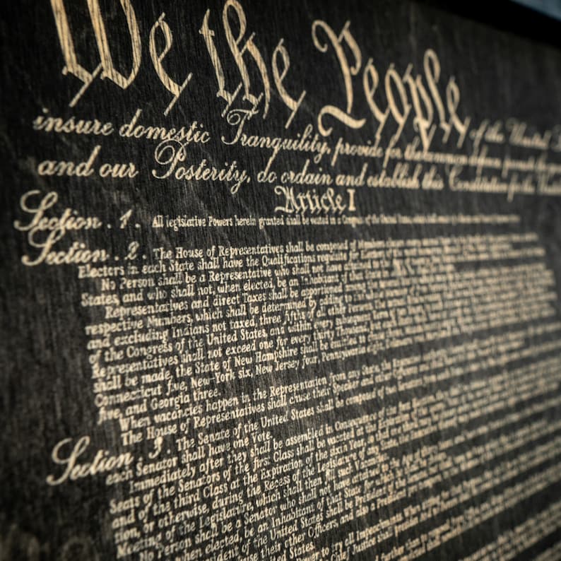 K&ouml;nnte beinhalten: Eine Nahaufnahme eines gerahmten Dokuments mit dem Text "We the People" und den ersten Abschnitten der Verfassung der Vereinigten Staaten. Das Dokument ist auf einem dunklen Hintergrund mit wei&szlig;em Text gedruckt.