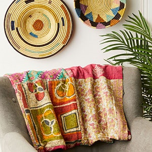 以下が含まれることがあります： ピンク、オレンジ、グリーン、ブルーの色合いのパッチワークデザインが施されたカラフルなカンタキルトスロー。キルトは、後ろに織られた壁掛けのあるグレーのソファにかけられています。