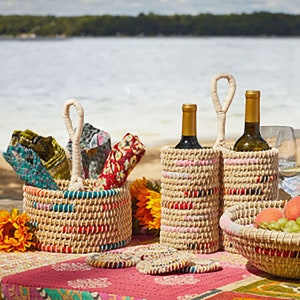 以下が含まれることがあります： ハンドル付きの織りバスケットが2つ、1つはワインボトル、もう1つはカラフルな布が入っています。バスケットはカラフルなストライプのデザインで、花柄のテーブルクロスのあるテーブルの上に置かれています。