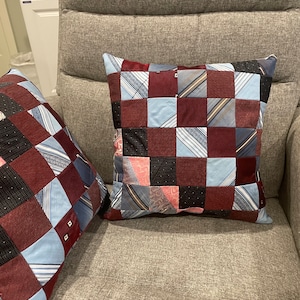 May include: Two square throw pillows with patchwork designs. The pillows are made from squares of various fabrics, including burgundy, light blue, and patterned materials. The pillows are on a gray upholstered chair.
