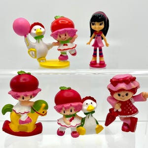 Może przedstawiać: Kolekcja kolorowych miniaturowych figurek z motywem truskawki. Figurki obejmują postacie z czerwonymi kapeluszami w kształcie truskawek, gęś trzymającą różowy balon i dziewczynkę z czarnymi włosami. Figurki są wykonane z tworzywa sztucznego.