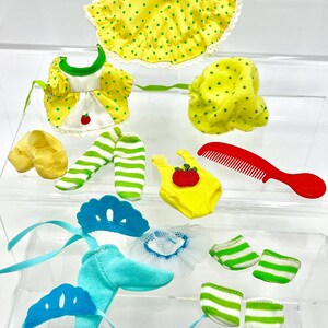 May include: A collection of doll clothes and accessories in various colors. Includes a yellow hat with green polka dots, a yellow jacket with a white dress, green and white striped leggings, and a red comb.