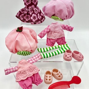 May include: A collection of doll clothes and accessories in shades of pink and red. Includes a floral dress, hats, shirts, pants, socks, shoes, and a red comb. The outfits have a vintage aesthetic.