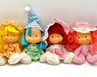 May include: Four vintage dolls with smiling faces, each with different coloured hair and outfits. The dolls have yellow, blue, red, and pink hair, and wear matching hats and pyjamas. The dolls are sitting on a white surface.
