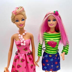 Puede incluir: Dos muñecas de moda, una rubia y rosa, la otra rosa. Una lleva un vestido rosa con estampado de fresas y un collar de perlas. La otra tiene pelo rosa, una camiseta verde a rayas con un gráfico de frambuesa y una falda vaquera azul.