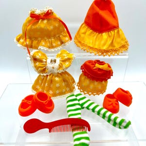 May include: A collection of doll clothes and accessories, including yellow floral dresses, orange hats, red shoes, green and white striped socks, and a red comb. The dresses have white lace trim and red bows. The accessories are arranged on a clear surface.
