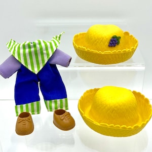 Puede incluir: Un conjunto de ropa de muñeca que incluye una camisa morada, pantalones azules y zapatos marrones. El conjunto incluye un babero a rayas verdes y blancas y dos sombreros amarillos. Un sombrero tiene un adorno de uvas moradas.