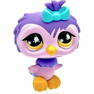 May include: A purple and pink owl figurine with a teal bow on its head. The owl has large eyes with starburst details, a yellow beak, and yellow feet. The figurine is a collectible toy.