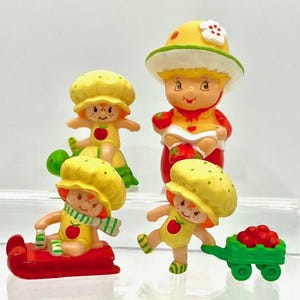 May include: Four vintage Strawberry Shortcake figurines. Each figure is wearing a yellow hat and outfit with a red apple emblem. One figure is on a red sled, another is pushing a green cart filled with red berries, and another is riding a green turtle.