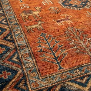 Puede incluir: Primer plano de una alfombra detallada de tonos anaranjados con un borde geométrico. La alfombra presenta diseños de camellos, árboles y geométricos en tonos azules, beige y marrón. La textura parece suave y acogedora.