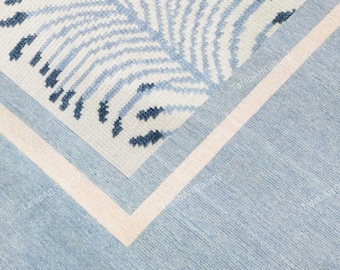 May include: A close-up of a blue and white patterned rug. The rug features a central design of a stylized leaf in shades of blue on a white background. A light pink border frames the design, set against a light blue background.