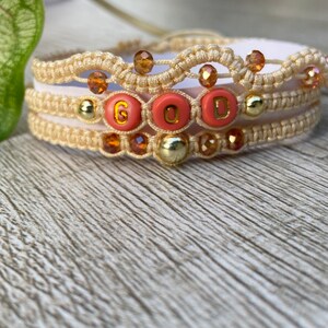 May include: Three layered bracelets with beige string, gold beads, and orange glass beads. The middle layer has coral colored beads with the word "GOOD" spelled out in gold letters.