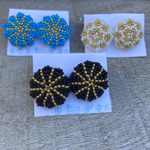 May include: Three pairs of beaded earrings. The first pair is blue with gold accents, the second pair is white with gold accents, and the third pair is black with gold accents.