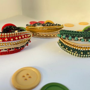 May include: Three bracelets made of woven threads and beads. The bracelets are in red, green, and gold colors. The bracelets are on a white surface.
