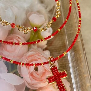 May include: Three necklaces with red and gold accents. The top necklace is gold with red beads. The middle necklace is red beaded with gold accents. The bottom necklace has a red cross pendant with gold trim and red beads.
