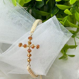 May include: A gold-toned cross bracelet with amber-colored beads. The cross is the focal point, with a woven cord band and adjustable string closure. The background includes white fabric and green foliage.