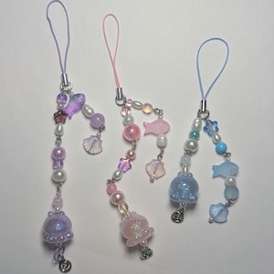 May include: Three colorful phone charms with a sea theme. Each charm features a different color scheme: purple, pink, and blue. Each charm has a variety of beads, including pearls, stars, seashells, and fish. The charms are attached to a string loop.