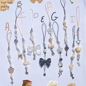 May include: A collection of decorative phone charms with various designs, including bows, stars, and floral elements. The charms feature beads, pearls, and metallic accents. The color palette includes white, cream, and silver.