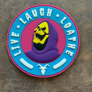 May include: A circular patch featuring a central purple-hooded yellow skull character on a pink background. A bright blue border surrounds the center, displaying the white text LIVE LAUGH LOATHE and a small white ram skull icon at the bottom.