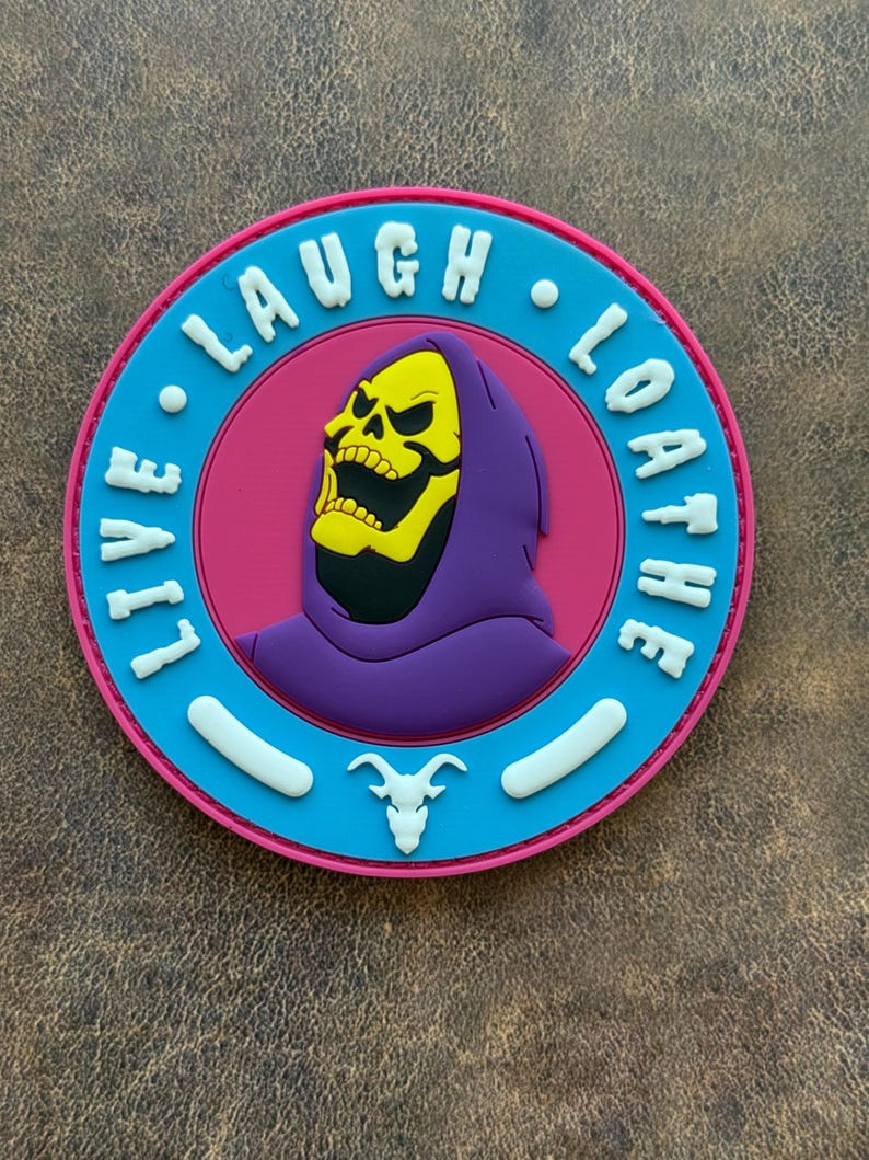 Op de afbeelding: Een ronde patch met een centraal geel schedelpersonage met een paarse capuchon op een roze achtergrond. Een felblauwe rand omringt het midden, met de witte tekst LIVE LAUGH LOATHE en een klein wit symbool van een ramschedel onderaan.