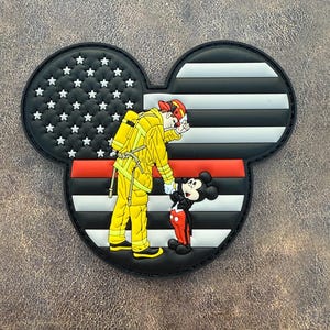 May include: A black, white, and red patch in the shape of Mickey Mouse's head. The left ear features a black background with white stars. The right ear has black and white stripes. A firefighter in yellow gear and Mickey Mouse are in the center.