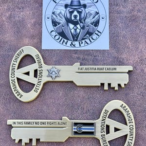 May include: Two brass-colored key-shaped coins with text and a badge. One coin reads "Arapahoe County Sheriff" and "Detentions Bureau". The other coin has the text "In This Family No One Fights Alone". A sticker with a bear wearing a hat is also present.