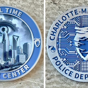 May include: Two silver challenge coins with blue accents. The first coin features a cityscape with an eye in the sky and the text "Real Time Crime Center". The second coin features a circuit board design with a stylized heart and the text "Charlotte-Mecklenburg Police Department".