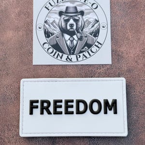 May include: Two patches on a brown surface. The top patch is square with a bear wearing a hat and sunglasses, smoking a pipe, and the text "FUZZ-FIVE-O COIN & PATCH". The bottom patch is rectangular with the word "FREEDOM".