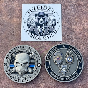 May include: Three collectible coins and a sticker. One coin features a skull with crossed weapons and the text "ARAPAHOE COUNTY NARCOTICS TEAM". The second coin displays police badges. The sticker has a bear wearing a suit and hat.