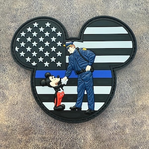 May include: A black, Mickey Mouse-shaped patch featuring an American flag design, a police officer, and Mickey Mouse shaking hands. The patch has a blue line and white stars. The officer is in a blue uniform.