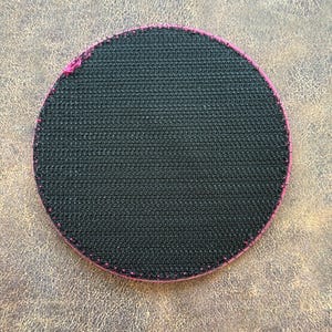 Op de afbeelding: Een cirkelvormige patch met een zwarte klittenbandachterkant. De rand van de patch is afgewerkt met een felroze gestikte boord, wat een duidelijk contrast vormt met het getextureerde zwarte oppervlak van het klittenbandmateriaal.