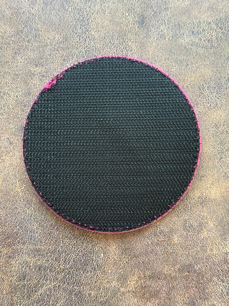 Op de afbeelding: Een cirkelvormige patch met een zwarte klittenbandachterkant. De rand van de patch is afgewerkt met een felroze gestikte boord, wat een duidelijk contrast vormt met het getextureerde zwarte oppervlak van het klittenbandmateriaal.