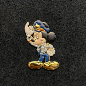 May include: A colorful enamel pin featuring Mickey Mouse in a police officer uniform. The pin has gold-colored shoes and accents, a blue hat with a gold star, and a white glove raised in a salute. The background is a dark gray fabric.
