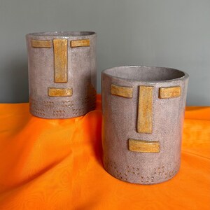 May include: Two gray ceramic planters with a face design. The face features a simple geometric design with a nose, eyes, and mouth. The planters are on a bright orange surface.