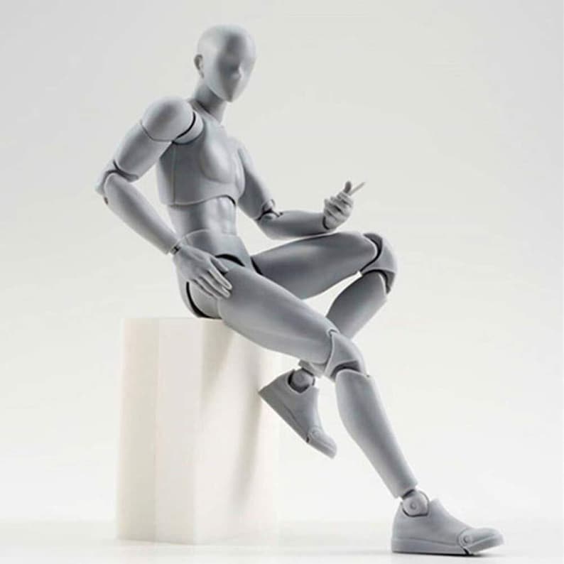 May include: A gray articulated figure sitting on a white cube. The figure is posed with one leg extended and the other bent at the knee. The figure has a neutral expression and is made of plastic.