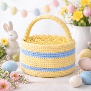 May include: A yellow and blue crochet Easter basket with a handle, filled with yellow straw. The basket has two horizontal blue stripes. Decorative Easter eggs and a ceramic bunny are in the background.