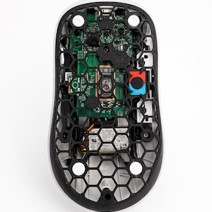 May include: A white and black computer mouse with a honeycomb design on the bottom, revealing the internal components. The mouse features a black honeycomb frame, a green circuit board, and red and blue buttons.