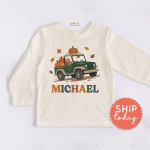 May include: A cream-colored long-sleeve shirt featuring a graphic of a green off-road vehicle carrying orange pumpkins on its roof and in the back. The name MICHAEL is printed in large, multi-colored letters below the vehicle illustration.