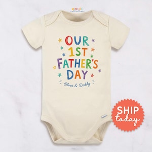 May include: Cream-colored baby onesie with the text "OUR 1ST FATHER'S DAY" in colorful letters and stars. The text "Oliver & Daddy" is below. A red and orange sticker with the words "SHIP today" is on the right.