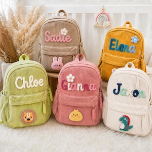 May include: Five corduroy backpacks in beige, yellow, light green, pink, and off-white. Each bag features a personalized name in embroidered letters, a small fabric flower, and a unique animal patch including a rabbit, lion, chick, and dinosaur.