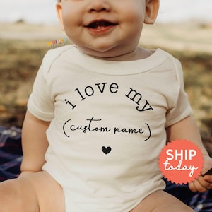 May include: Cream-colored baby bodysuit with the text "i love my (custom name)" in black script, a black heart, and a red "SHIP today" sticker. The bodysuit is short-sleeved.